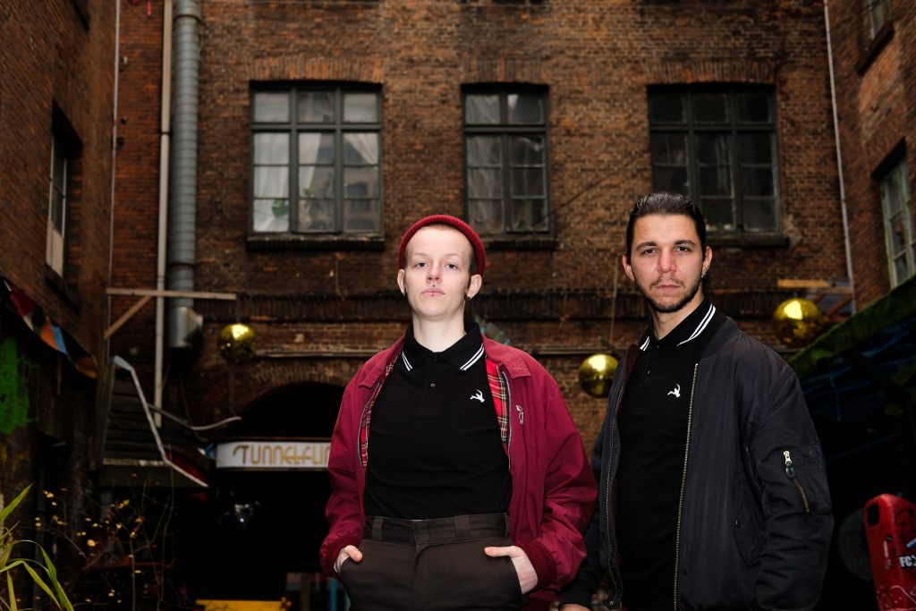 Zwei Queers mit Bomberjacke und Buttplug-und-Sichel-Polo-Shirt schauen herausforndernd in die Kamera. Im Hintergrund eine Backsteinfassade.